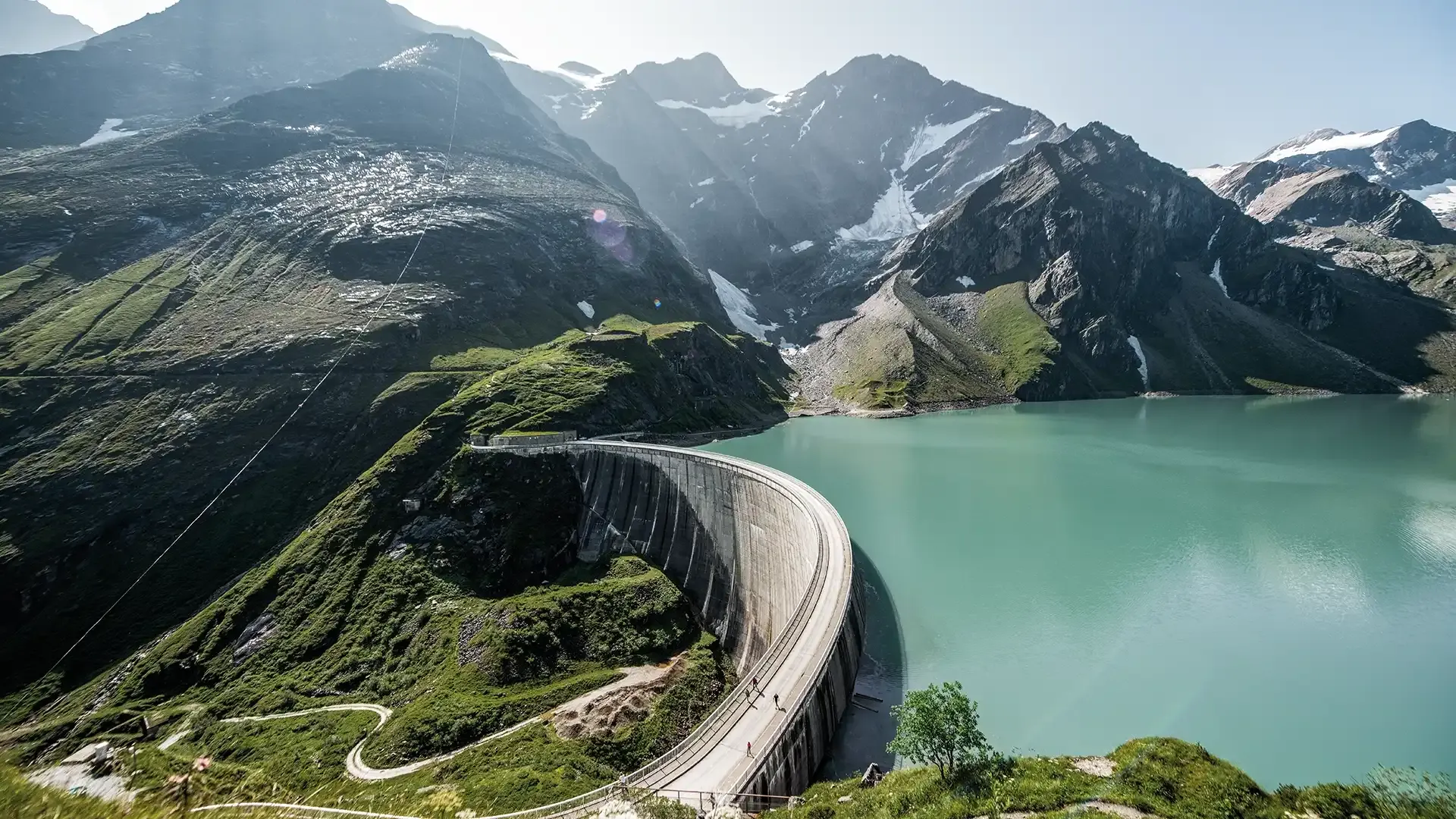 Eine Luftaufnahme von einem Staudamm in gebirgiger Landschaft, der Stausee ist klar und leuchtet türkis.