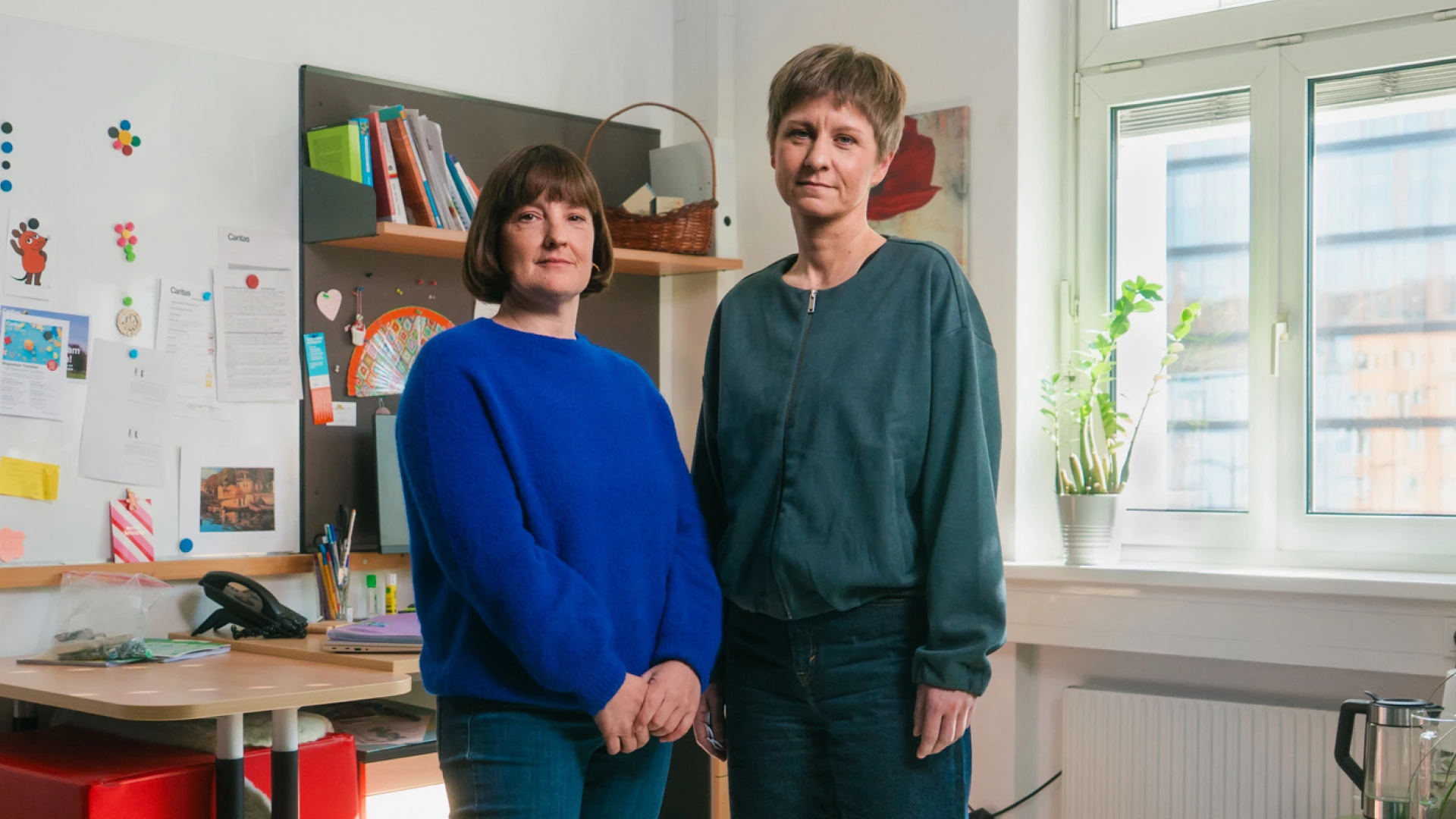 Zwei Frauen stehen nebeneinander. Die linke hat kürzeres braunes Haar und trägt einen blauen Pullover. Die rechte trägt ein petrolfarbenes Oberteil. Sie hat kurze blonde Haare.