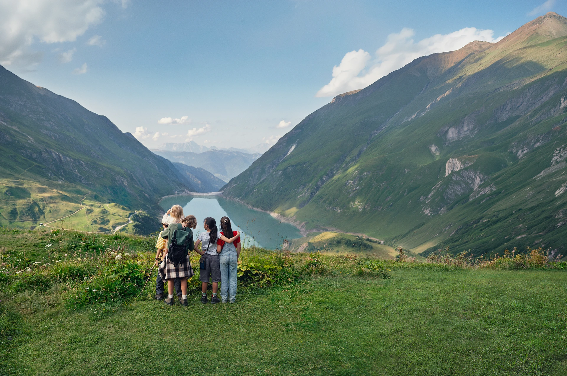 Fünf Kinder stehen auf einer Wiese und schauen in ein alpines Tal mit See zwischen grünen Bergen.